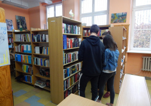 Wnętrze biblioteki szkolnej. Uczniowie przeglądają książki eksponowane na regale z nowościami.